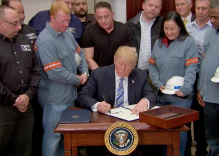 Donald Trump sentado a mesa, assinando documento com várias pessoas atrás dele.